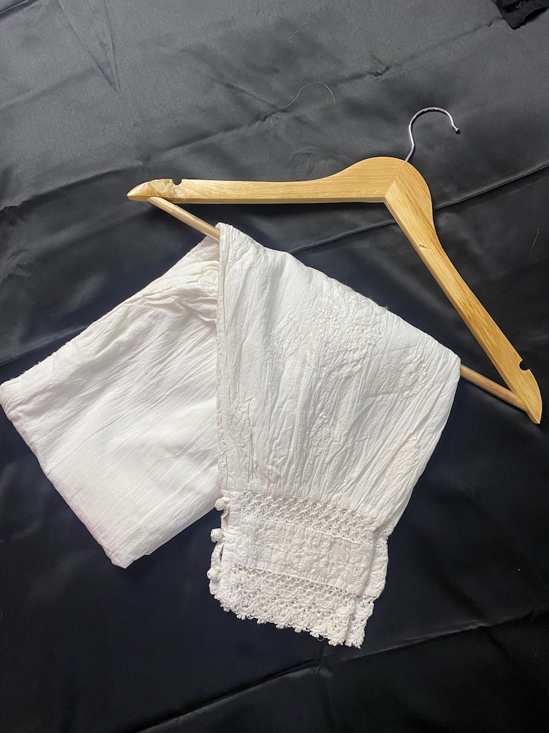 May include: White, textured fabric pants with a decorative lace trim at the ankle. The pants are displayed on a wooden hanger against a black background. The fabric appears to be lightweight and suitable for warm weather.