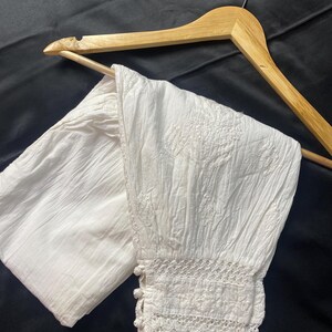 May include: White, textured pants with embroidered floral details and a lace trim at the ankle. The pants are hanging on a wooden hanger against a dark background. The pants appear to be made of a lightweight fabric.