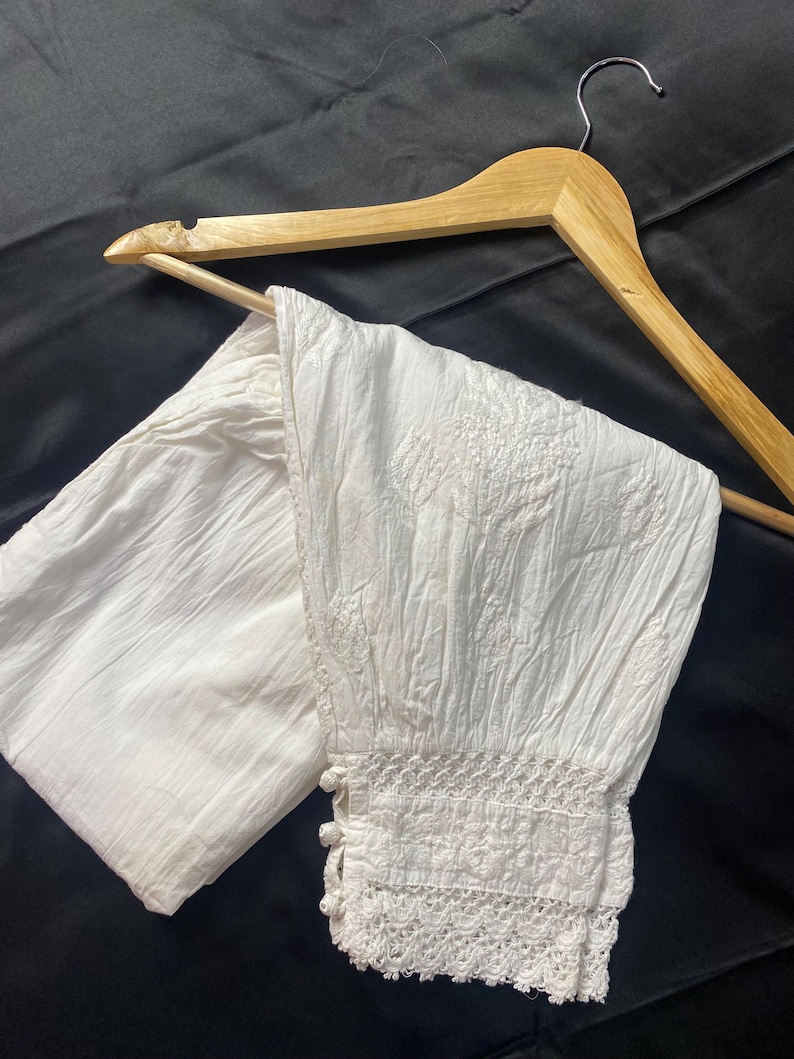 May include: White, textured pants with embroidered floral details and a lace trim at the ankle. The pants are hanging on a wooden hanger against a dark background. The pants appear to be made of a lightweight fabric.