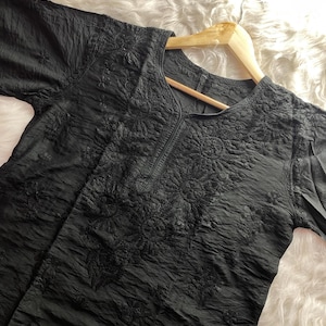 May include: A black tunic featuring a textured, crinkled fabric surface and intricate tonal floral embroidery around the neckline and chest area. The garment has long sleeves and a rounded neckline, displayed on a wooden hanger.
