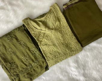 May include: An olive green embroidered three-piece outfit. The set includes a long-sleeved top with a V-neck, matching pants, and a scarf. The fabric has a textured appearance, and the embroidery is detailed. The items are arranged on a white, fluffy surface.