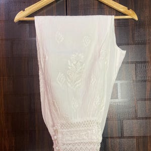 May include: White, embroidered pants hanging on a wooden hanger. The pants feature a detailed floral pattern and lace trim at the ankles. The fabric appears to be lightweight and suitable for warm weather.