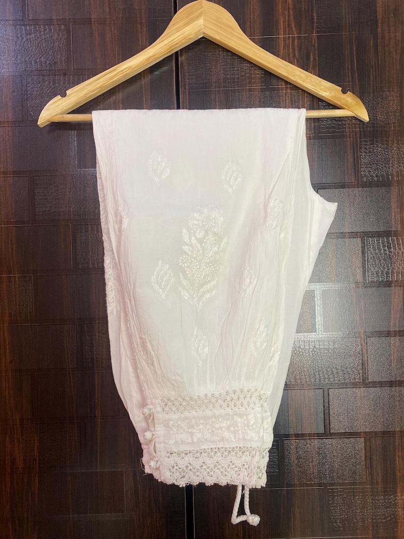 May include: White, embroidered pants hanging on a wooden hanger. The pants feature a detailed floral pattern and lace trim at the ankles. The fabric appears to be lightweight and suitable for warm weather.