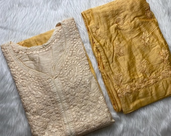 Puede incluir: Un conjunto de túnica bordada color crema y pantalón amarillo mostaza. La túnica presenta un bordado floral detallado y un cuello en V. El pantalón está doblado cuidadosamente junto a la túnica. El conjunto se muestra sobre una superficie blanca y esponjosa.