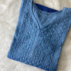 May include: A light blue embroidered tunic with a V-neck. The tunic features intricate floral patterns and a subtle ombre effect, transitioning from a lighter shade at the top to a deeper blue at the bottom. The garment is made of a soft, lightweight fabric.