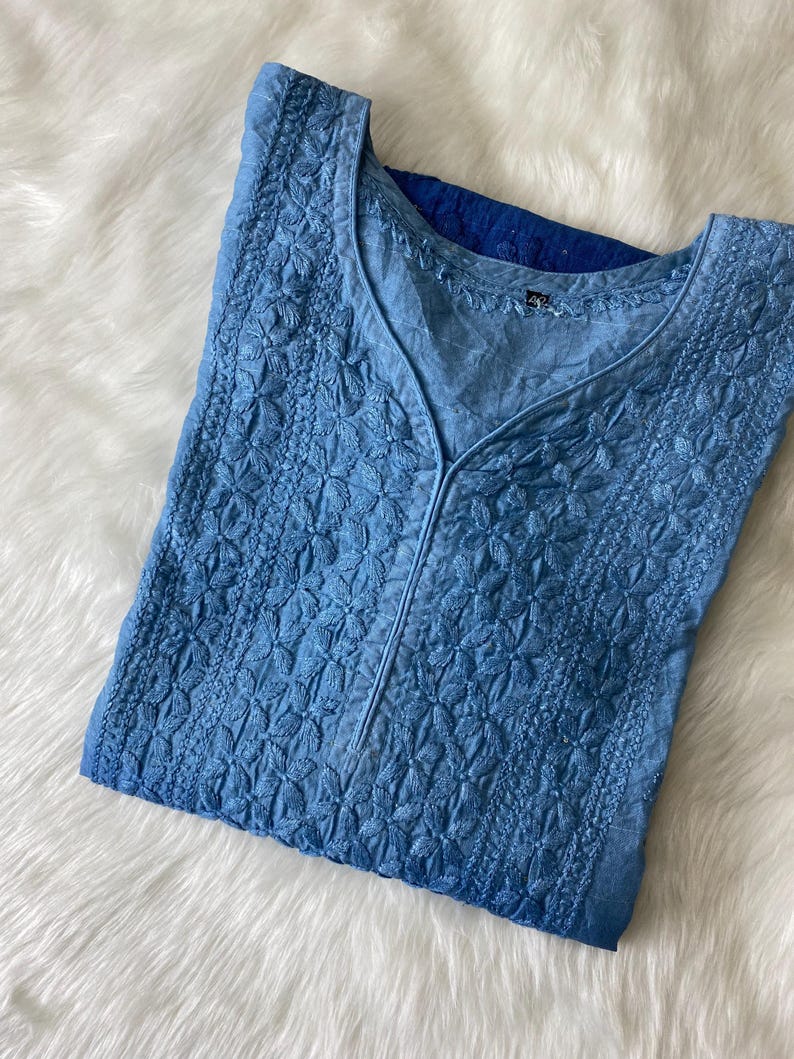 May include: A light blue embroidered tunic with a V-neck. The tunic features intricate floral patterns and a subtle ombre effect, transitioning from a lighter shade at the top to a deeper blue at the bottom. The garment is made of a soft, lightweight fabric.