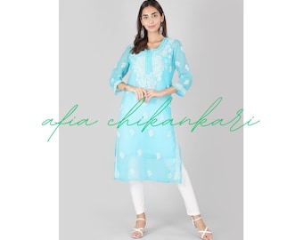 May include: A light blue, long-sleeved, embroidered tunic top with white embroidery. The top has a round neckline and a straight hem. The model is wearing white pants and white sandals.