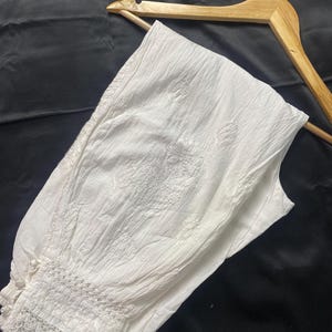 May include: White, embroidered cotton pants with a textured appearance. The pants feature a decorative lace trim at the ankle and are displayed on a wooden hanger against a dark background. The embroidery has a floral design.