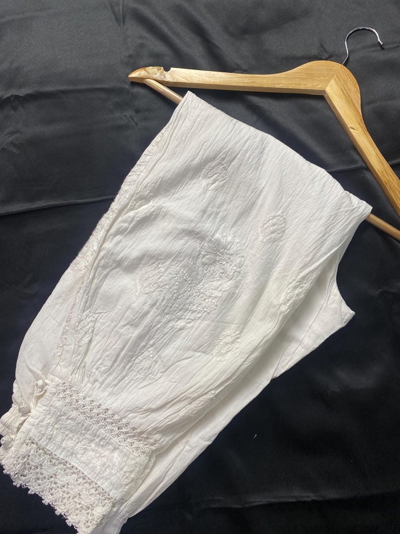 May include: White, embroidered cotton pants with a textured appearance. The pants feature a decorative lace trim at the ankle and are displayed on a wooden hanger against a dark background. The embroidery has a floral design.