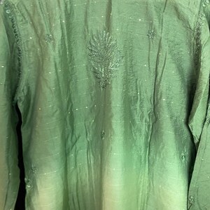 May include: A green long-sleeved tunic with intricate embroidery around the neckline and a central leaf-like design. The fabric has a subtle sheen and a gradient effect, transitioning from a darker green at the top to a lighter shade towards the bottom. Small, sparkling embellishments are scattered across the surface.