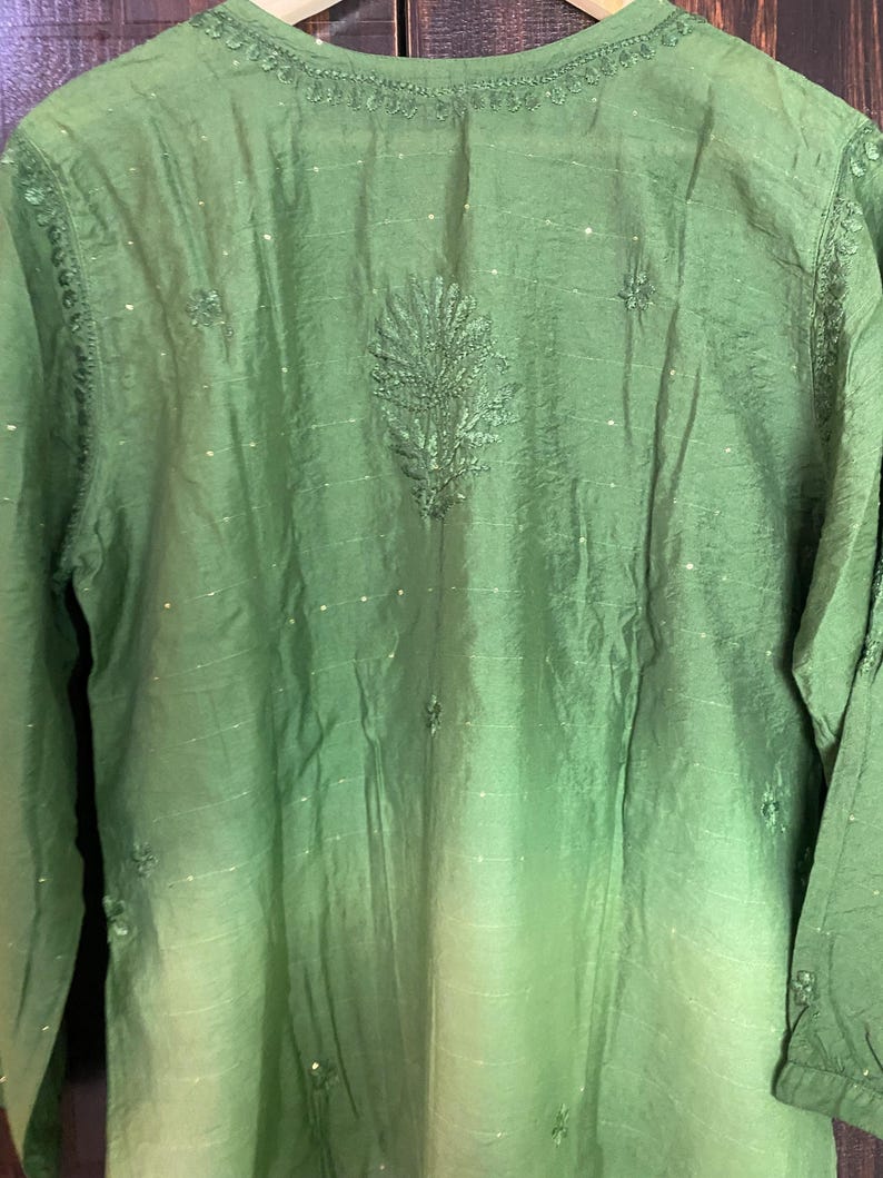 May include: A green long-sleeved tunic with intricate embroidery around the neckline and a central leaf-like design. The fabric has a subtle sheen and a gradient effect, transitioning from a darker green at the top to a lighter shade towards the bottom. Small, sparkling embellishments are scattered across the surface.