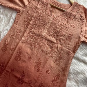 May include: A peach-coloured tunic with detailed floral embroidery. The long-sleeved garment features a V-neck and intricate patterns across the chest and sleeves. The fabric has a subtle sheen and a gradient effect, darkening towards the hem.