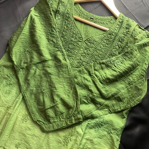 May include: A green tunic with long sleeves and a V-neck. The neckline and sleeves feature detailed embroidery. The tunic is hanging on a wooden hanger against a dark background. The fabric has a textured appearance.