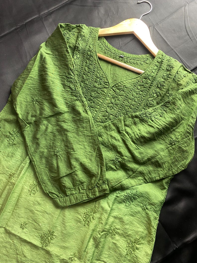May include: A green tunic with long sleeves and a V-neck. The neckline and sleeves feature detailed embroidery. The tunic is hanging on a wooden hanger against a dark background. The fabric has a textured appearance.