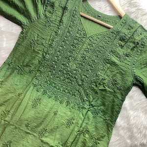 May include: A green tunic with intricate embroidery around the neckline and bodice. The fabric has a textured appearance and a subtle sheen. The tunic is displayed on a wooden hanger.
