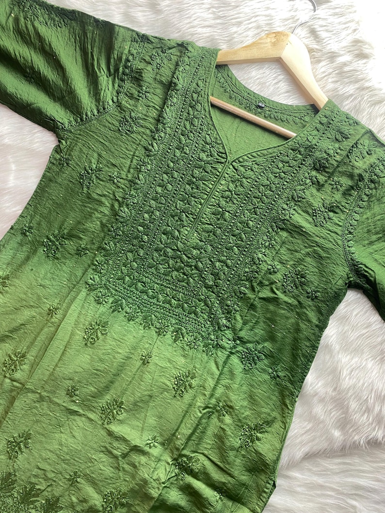 May include: A green tunic with intricate embroidery around the neckline and bodice. The fabric has a textured appearance and a subtle sheen. The tunic is displayed on a wooden hanger.
