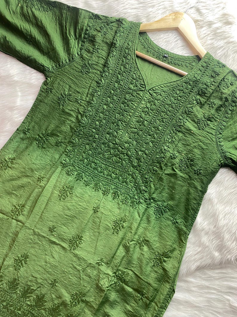 May include: A green tunic with intricate embroidery around the neckline and sleeves. The fabric has a textured appearance and a subtle sheen. The tunic is displayed on a wooden hanger, showcasing its design and details.
