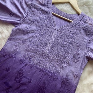 Può includere: Una tunica sfumata lavanda e viola con ricami floreali intricati. Il top a maniche corte presenta uno scollo a V ed è esposto su una gruccia di legno. Il tessuto appare leggero e leggermente trasparente.