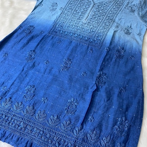 May include: A blue tunic with a V-neck and short sleeves. The fabric has a gradient effect, transitioning from light blue at the top to a deep blue at the bottom. Intricate floral embroidery adorns the neckline and hem.