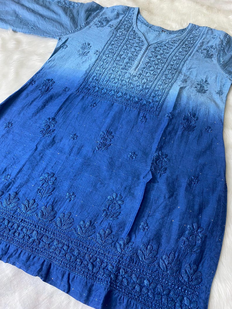 May include: A blue tunic with a V-neck and short sleeves. The fabric has a gradient effect, transitioning from light blue at the top to a deep blue at the bottom. Intricate floral embroidery adorns the neckline and hem.