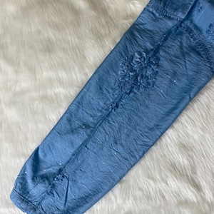 May include: A light blue, long-sleeved garment with intricate floral embroidery. The fabric has a crinkled texture and is embellished with small, sparkling accents. The sleeve is laid on a white, fluffy surface.