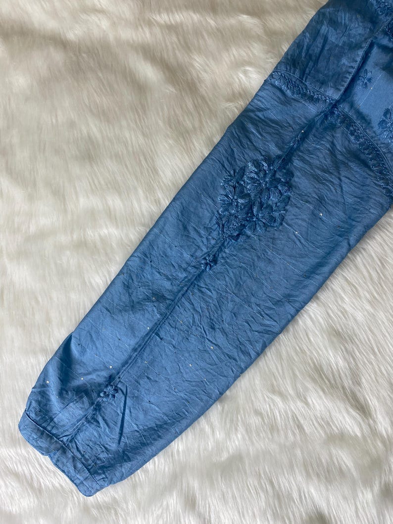 May include: A light blue, long-sleeved garment with intricate floral embroidery. The fabric has a crinkled texture and is embellished with small, sparkling accents. The sleeve is laid on a white, fluffy surface.