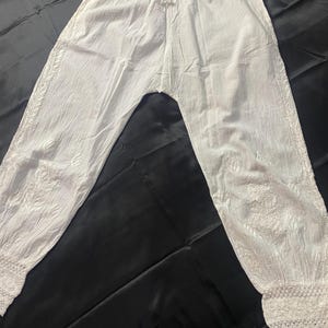 May include: White, lightweight pants with an elastic waistband and drawstring. The pants feature embroidered detailing along the sides and a delicate lace trim at the ankles. The fabric appears to be slightly textured.