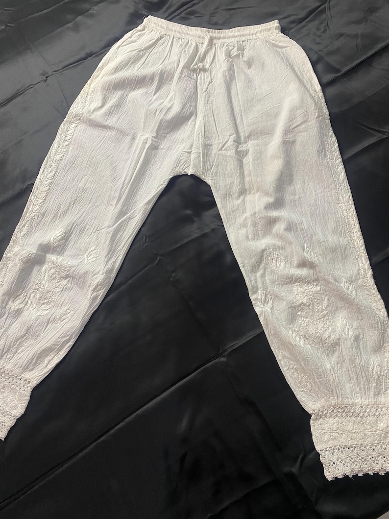 May include: White, lightweight pants with an elastic waistband and drawstring. The pants feature embroidered detailing along the sides and a delicate lace trim at the ankles. The fabric appears to be slightly textured.