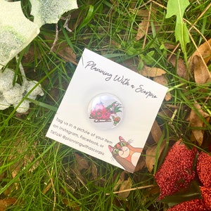 May include: A white card with the text "Planning With a Scorpiu" and a small round button with a cartoon image of a red sleigh with a Christmas tree and a cat. The text on the card says "Tag us in a picture of your item on Instagram, Facebook, or TikTok @planningwithascorpio".