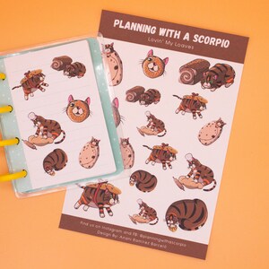 May include: A planner and sticker sheet featuring cat illustrations. The stickers and planner pages have a light blue background with cat and bread-themed designs. The text "PLANNING WITH A SCORPIO" is visible on the sticker sheet.