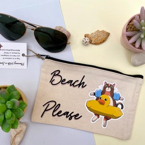May include: A beige canvas pouch with a black zipper and the text "Beach Please" in black cursive lettering. A cartoon cat wearing blue armbands is riding a yellow rubber ducky.