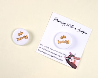 Könnte beinhalten: Zwei weiße runde Pins mit dem Text "oh honey" in goldener Schrift. Die Pins befinden sich auf einem weißen Hintergrund mit einer Karte, auf der "Planning With a Scorpio" steht und ein Comic-Katze abgebildet ist.