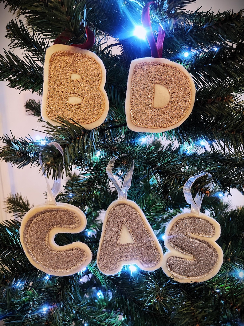 K&ouml;nnte beinhalten: Goldene und silberne Glitzer-Filzbuchstaben-Ornamente, die den Namen "BD CAS" buchstabieren. Die Ornamente h&auml;ngen an einem gr&uuml;nen Weihnachtsbaum.