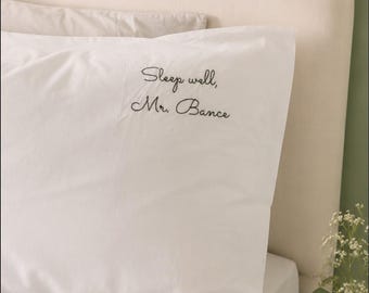 Könnte beinhalten: Ein weißer Kissenbezug mit schwarzem, gesticktem Schreibschrifttext, der "Sleep well, Mr. Bance" lautet und sich in der oberen rechten Ecke befindet. Der Stoff ist glatt und schlicht und bildet einen Kontrast zur gemusterten Bettwäsche darunter.
