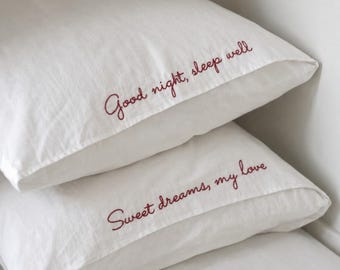 Könnte beinhalten: Zwei weiße rechteckige Kissenbezüge, die übereinander gestapelt sind. Der obere Kissenbezug zeigt den gestickten dunkelroten kursiven Text "Good night, sleep well" und der untere Kissenbezug den gestickten dunkelroten Text "Sweet dreams, my love".