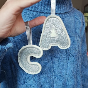 K&ouml;nnte beinhalten: Zwei Filz-Anh&auml;nger mit silbernen Glitzerbuchstaben, die das Wort "CA" ergeben. Die Anh&auml;nger sind wei&szlig; mit silbernem Glitzerfinish und h&auml;ngen an einem wei&szlig;en Band.