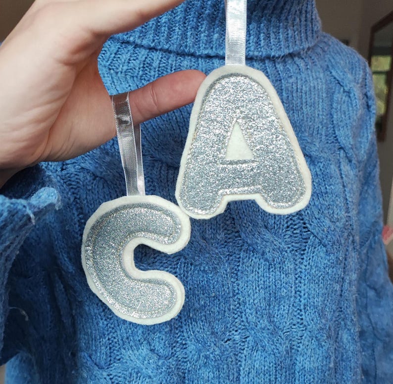 K&ouml;nnte beinhalten: Zwei Filz-Anh&auml;nger mit silbernen Glitzerbuchstaben, die das Wort "CA" ergeben. Die Anh&auml;nger sind wei&szlig; mit silbernem Glitzerfinish und h&auml;ngen an einem wei&szlig;en Band.