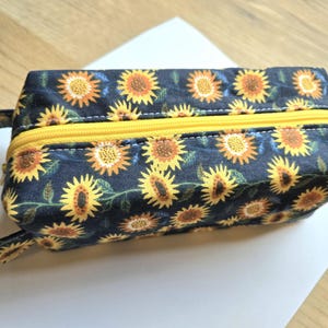 May include: A rectangular zippered pouch featuring a dark navy blue fabric base with a repeating pattern of yellow and orange sunflowers and green leaves. The pouch includes a bright yellow zipper closure and a matching fabric carrying strap.