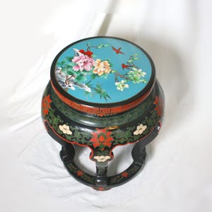 May include: A round, black, decorative table with a turquoise top featuring a floral design with pink, yellow, and white flowers, and red birds. The table has a red and green border and floral accents.