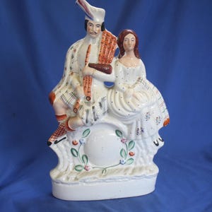 May include: A white ceramic figurine depicting a man in traditional Scottish attire playing a musical instrument, seated next to a woman in a white dress. The figurine has a floral and leaf design around the base.