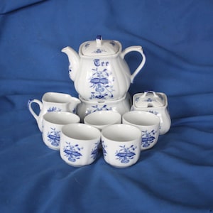 May include: A white porcelain tea set with blue floral designs. The set includes a teapot with the word "Tee", a creamer, a sugar bowl, and six teacups. The tea set is arranged on a blue surface.