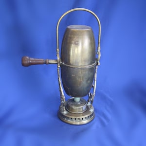 May include: Antique brass coffee maker with a wooden handle and a curved metal handle. The coffee maker has an oval-shaped body and sits on a round base with small holes. The background is a solid blue.