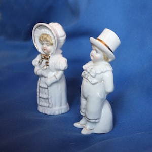 May include: A pair of vintage porcelain figurines of a girl and boy dressed in white with gold accents. The girl is wearing a bonnet and the boy is wearing a top hat.