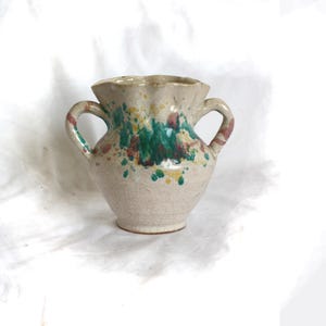 May include: A small, white ceramic vase with two handles. The vase has a scalloped rim and is decorated with green, yellow, and pink splatters.