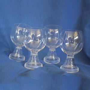 May include: Four clear, stemmed glasses with rounded bowls and decorative bases. The glasses are made of transparent glass, reflecting light and the blue background. The bases have a detailed, ridged design. The glasses are arranged on a blue surface.