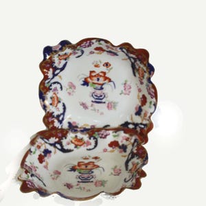 May include: Two antique, scalloped-edge porcelain bowls. The bowls feature a white background with floral and foliate designs in red, blue, and orange. The rims are trimmed in a dark red and blue. The bowls are stacked on top of each other.