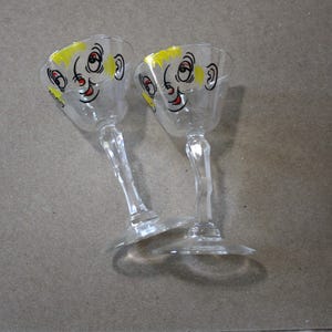 May include: Two clear glass goblets with a whimsical face design. The faces have yellow hair, black eyes, and red smiles. The goblets have a stem and a round base, suitable for serving beverages.