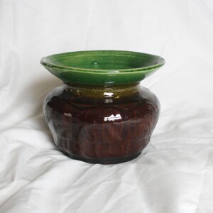 May include: A handcrafted ceramic vase with a wide, green rim and a bulbous, textured body. The vase transitions from a dark brown base to a lighter brown midsection, with a glossy finish. The vase is a decorative item.