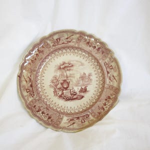 May include: A decorative plate with a scalloped edge. The plate features a detailed design in shades of brown and cream, depicting a landscape scene with figures and architectural elements. The plate is set against a white background.