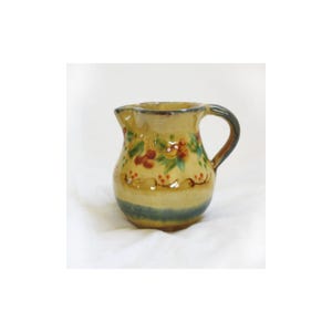 May include: A small, hand-painted ceramic pitcher with a curved handle and spout. The pitcher features a yellow base with a green band at the bottom and is decorated with red and green floral designs. The handle is dark brown.